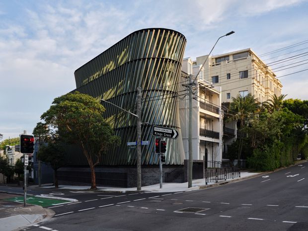 Street-level exterior view of a distinctive modern multi-storey building with a curved, sculptural facade wrapped in vertical dark fins and reflective glazing. It stands at a landscaped urban intersection beside older residential or commercial buildings, with traffic lights, road markings, utility wires, and directional signage visible.