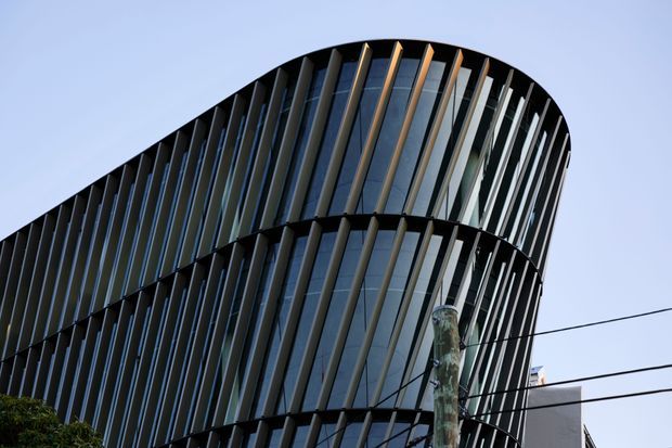 Upward-looking exterior view of a contemporary multi-storey building with a strongly curved facade. The dark glazed envelope is articulated by dense vertical fins or louvers, creating a rhythmic sculptural pattern around the rounded corners. Utility wires and a street-side pole cross the foreground, with pale blue sky behind.