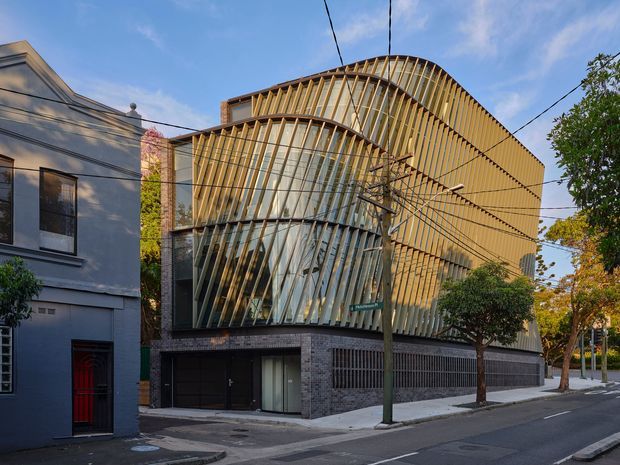 Street-level exterior view of a contemporary multi-storey urban building. The facade combines dark brick at the lower levels with extensive curved glazing and a prominent sculptural screen of closely spaced timber-toned vertical fins that wrap over the upper volume. Adjacent low-rise residential buildings, street trees, overhead utility wires and a paved road frame the building.