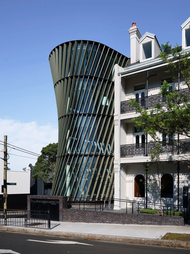 Street-level exterior view showing a striking contemporary multi-storey building with a curved, twisting form, extensive glazing, and repeated dark vertical fins beside a restored ornate white heritage terrace building with balconies, decorative railings, arched openings, and gabled dormers. The buildings sit directly on an urban street with a brick retaining wall, pedestrian ramp, fencing, and overhead utility lines under a clear blue sky.
