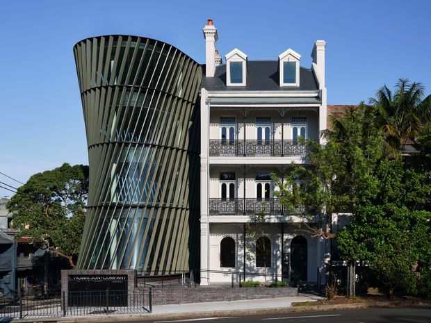 Street-facing view of a heritage-style multi-storey townhouse or terrace building with ornate balconies, arched openings, decorative window surrounds, dormer windows and chimneys, directly adjoining a striking contemporary addition clad in vertical curved fins and extensive glazing. Mature street trees and palms partially frame the property.