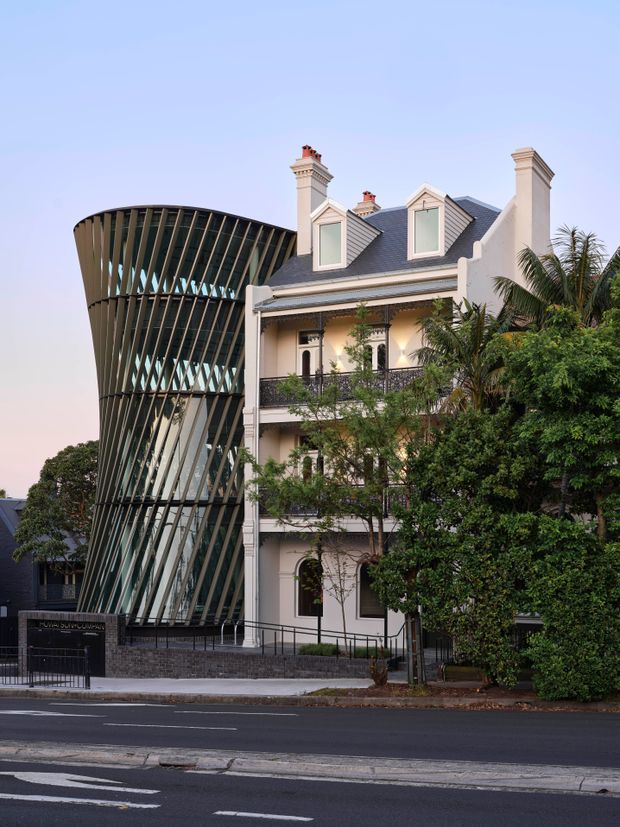 Street-facing exterior view of a heritage-style multi-storey building with a steep dark slate roof, ornate chimneys, dormer windows, decorative balcony railings and arched openings. A striking contemporary glazed and curved vertical-louvered addition rises alongside it, creating a strong contrast between traditional and modern architecture. Mature trees partially screen the facade, with a paved street in the foreground.