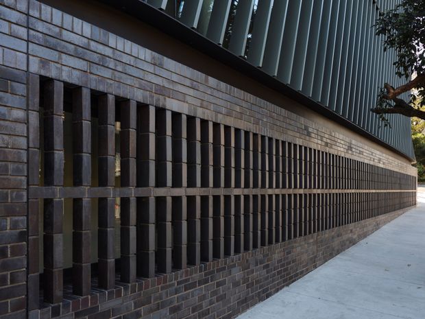 Close-up exterior view of a contemporary building facade featuring dark brick masonry, deep vertical brick fins forming a repetitive screened pattern, and a ribbed dark metal upper facade. A concrete pedestrian path runs alongside the wall.