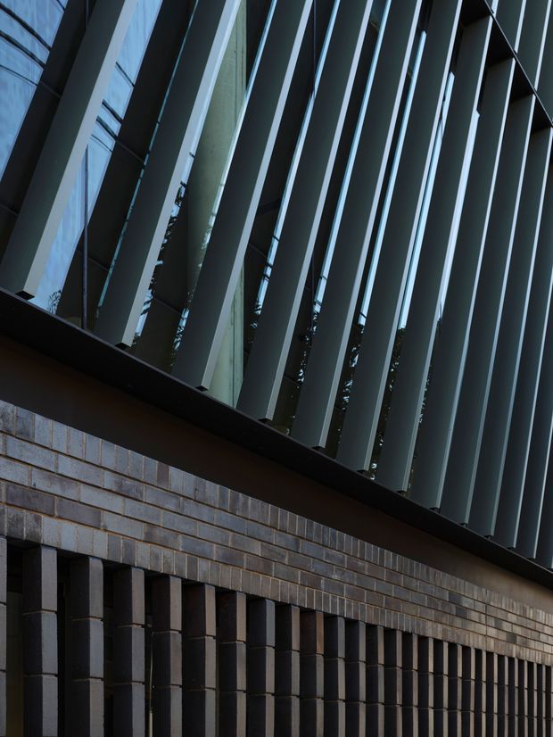 Close-up architectural exterior detail showing a dark brick facade with projecting vertical brick piers and a dramatic upper screen of repeated angled metal fins over glazing. The composition is geometric and linear, with a dark, restrained contemporary palette and glimpses of trees and sky behind the screen.