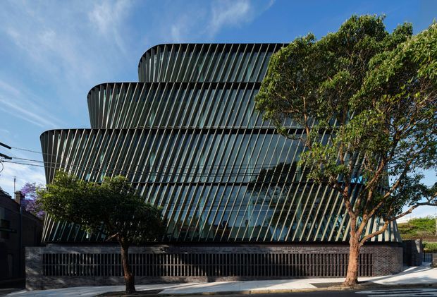 Street-level exterior view of a contemporary multi-storey commercial building with a sculptural stepped form and sweeping curved volumes. The facade is largely glazed and screened by dense vertical fins, creating a strong repetitive rhythm. A dark perimeter fence, mature street trees, sidewalk, and overhead utility lines frame the building.
