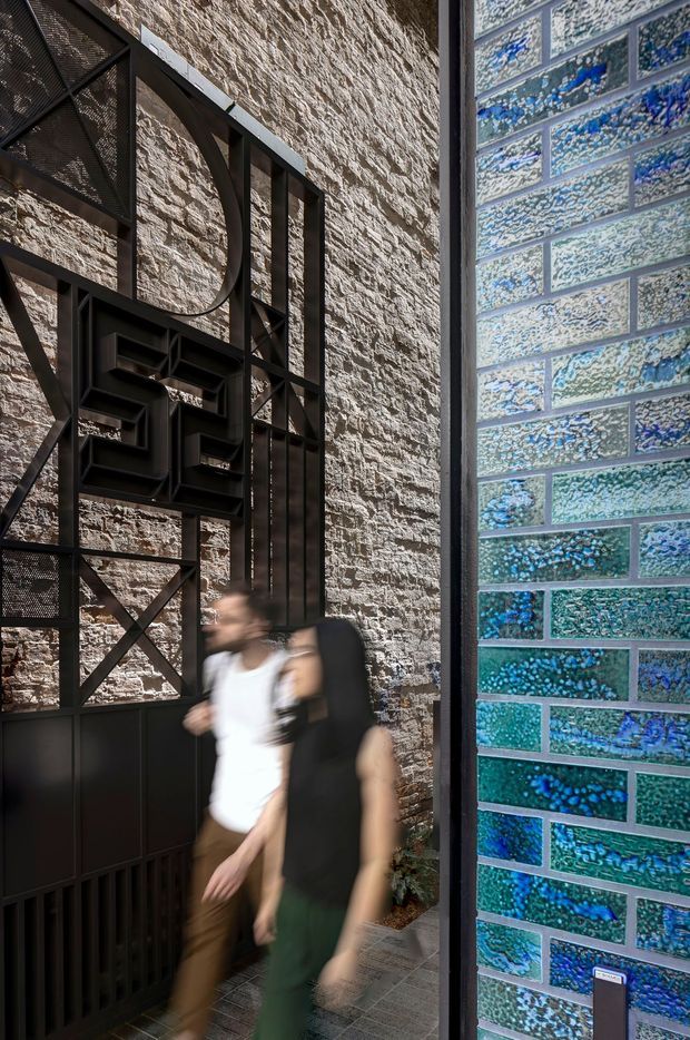 A narrow architectural passage or entry space with a heavily textured natural-stone wall, dark metal structural screening and sculptural geometric artwork, and a striking wall of blue and turquoise glazed brick tiles. Two blurred people are walking through the space. The composition highlights contrasting rugged stone, black metalwork, and iridescent blue tile surfaces in a contemporary, gallery-like setting.