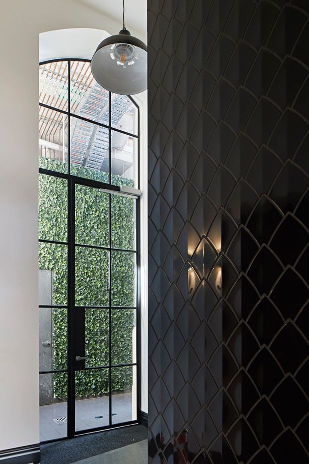 A tall, refined interior entry space with a black-framed, floor-to-ceiling glazed door opening to a small planted courtyard. The right wall is covered in a dramatic glossy black geometric fish-scale or overlapping-arch feature surface, complemented by a small wall sconce. A sculptural glass pendant hangs near the high ceiling, while the pale plaster wall and dark floor create strong tonal contrast.