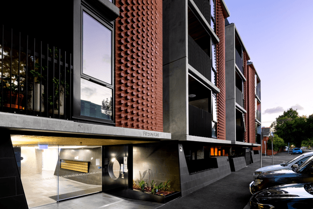 Street-level exterior view of a contemporary multi-storey apartment building. The facade combines red brick breeze-block screens, dark concrete or metal panels, recessed balconies with black balustrades, large windows, and a glazed illuminated entry lobby. Landscaped planter beds and parked cars line the urban frontage.