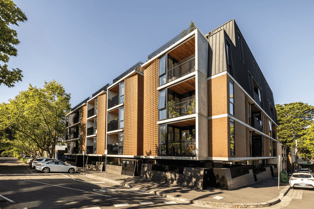 Exterior view of a contemporary multi-storey apartment building on a corner site. The facade combines exposed red brick panels, raw concrete structural elements, dark metal cladding, black balcony railings, and timber-lined balcony ceilings. The building is elevated above a partially open ground level, with adjacent paved parking and mature street trees.