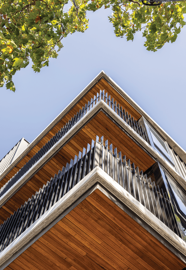 Upward-looking architectural photograph of a multi-storey building with strongly projecting, stacked balconies. The soffits are clad in warm timber boards, framed by raw concrete edges and lined with slim vertical metal balustrades. A clear blue sky and leafy tree canopy surround the building, emphasizing its geometric, cantilevered form.