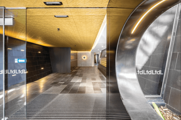 A polished commercial lobby or entrance with a dramatic curved metallic portal framing a glazed doorway. The interior features glossy black tiled walls, patterned gold ceiling panels with recessed lighting, grey and beige stone-look floor tiles, and a ribbed entrance mat. Reflections and signage are visible on the glass.
