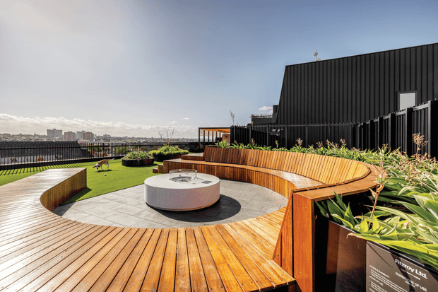 Rooftop terrace with expansive timber decking, built-in curved timber bench seating, a circular paved fire-pit or table area, raised planters, and artificial lawn. A dark ribbed-clad building volume borders the terrace, with city buildings and a distant coastal or urban horizon beyond the railing. Bright contemporary outdoor space with warm timber and lush planting.