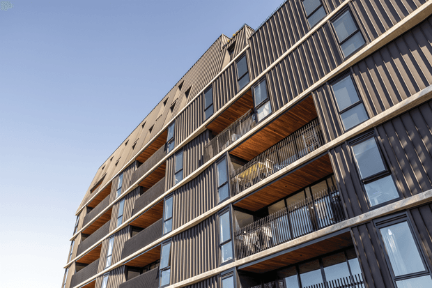 Upward-looking exterior view of a multi-storey apartment building with a strongly repetitive facade. The building combines dark vertically ribbed cladding, concrete balcony slabs and structural bands, large dark-framed windows, and recessed balconies with warm timber soffits and black metal balustrades. Clear blue sky forms the background.