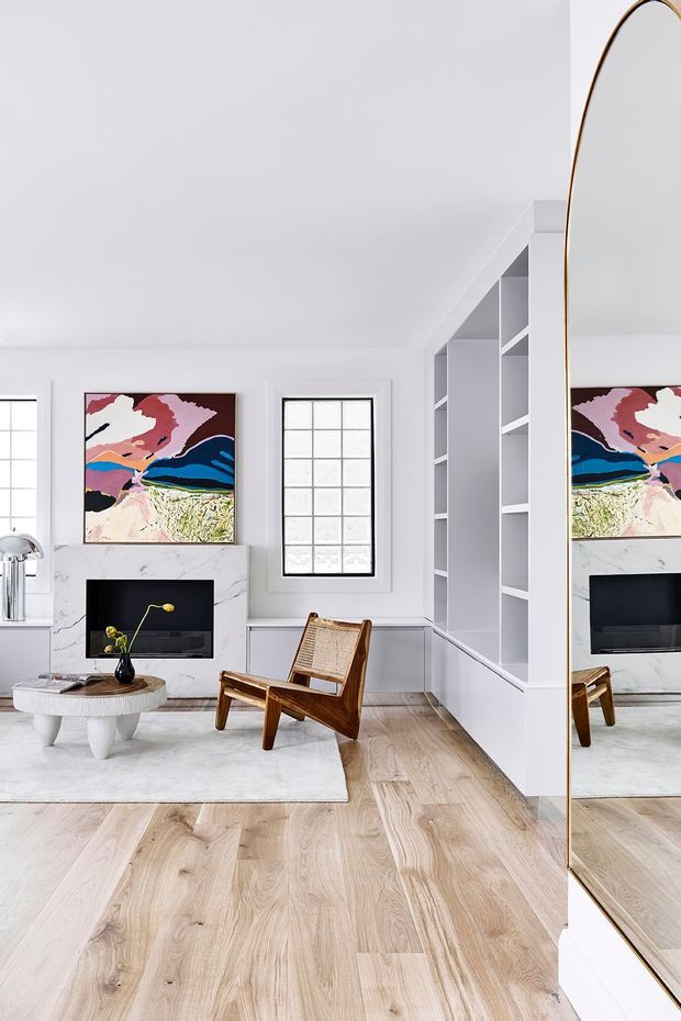Bright, refined living room with white walls and built-in open shelving surrounding a marble fireplace. A sculptural white coffee table, wooden lounge chair, colourful abstract artwork, pale timber flooring and a large arched-edge mirror create a contemporary, gallery-like interior.