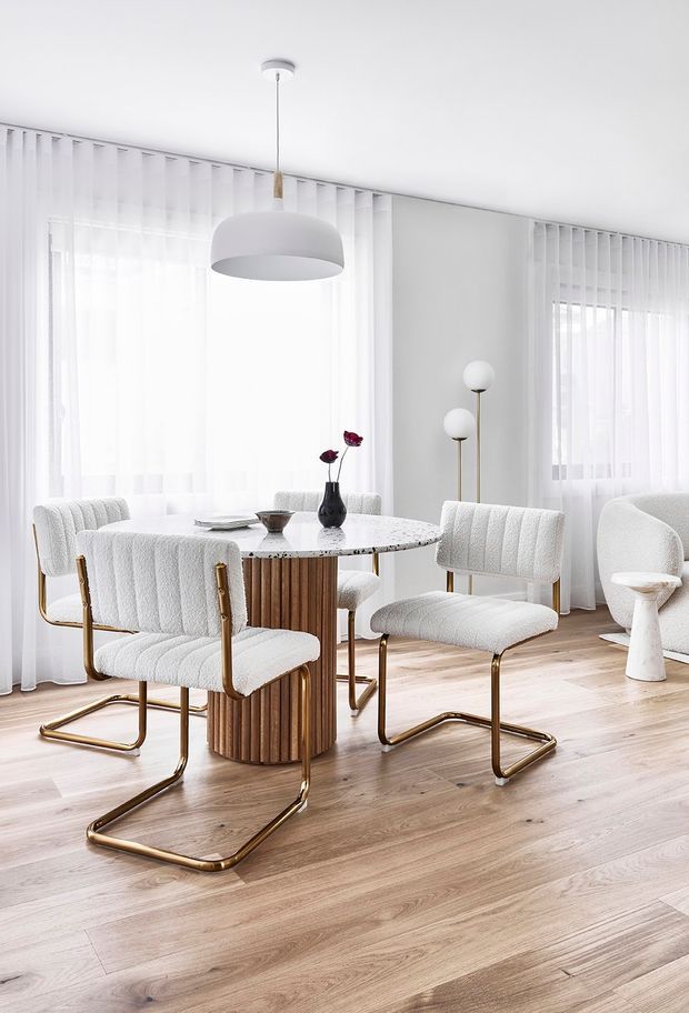 Bright, refined dining area with a round marble-top table, fluted timber pedestal base, and four upholstered cantilever dining chairs with brass-toned metal frames. Sheer curtains and vertical blinds span the windows, while sculptural pendant and floor lamps complement the pale timber flooring and soft white palette.