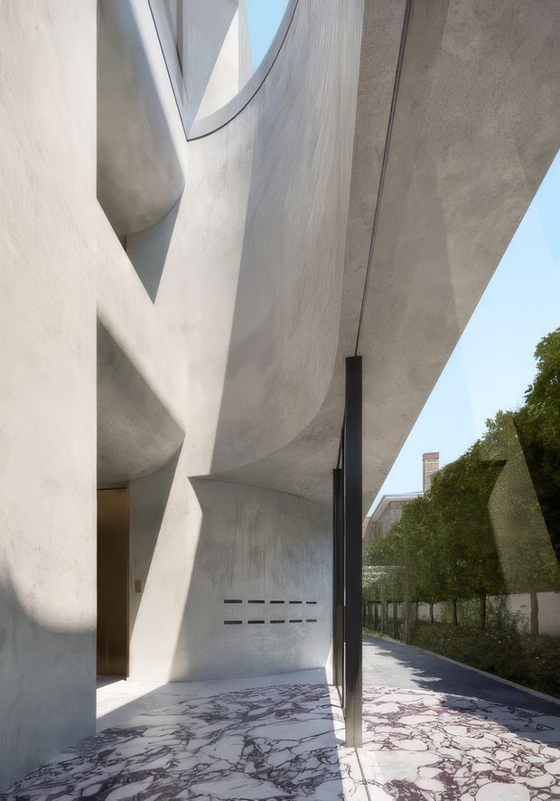 A dramatic contemporary residential entrance beneath sweeping sculptural concrete forms. The facade and deep overhanging canopy are finished in pale textured plaster or concrete, with a tall dark pivot door and narrow black vertical element. The ground is clad in striking white stone with dark irregular veining, while a landscaped hedge and neighbouring residential context appear along the walkway. Strong sunlight and curved geometry create a sculptural, high-contrast architectural composition.