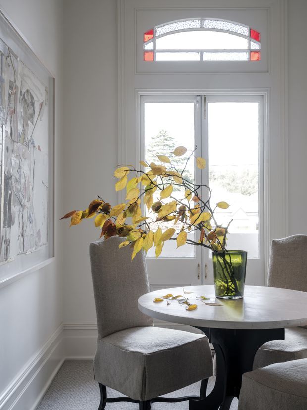 Bright, refined dining area with a round white dining table, upholstered grey slipcovered chairs, and a tall glass vase containing autumn branches. The room features pale moulded walls, traditional trim, a large artwork, carpeted flooring, and a tall glazed door/window with an arched decorative stained-glass transom in red and patterned glass.