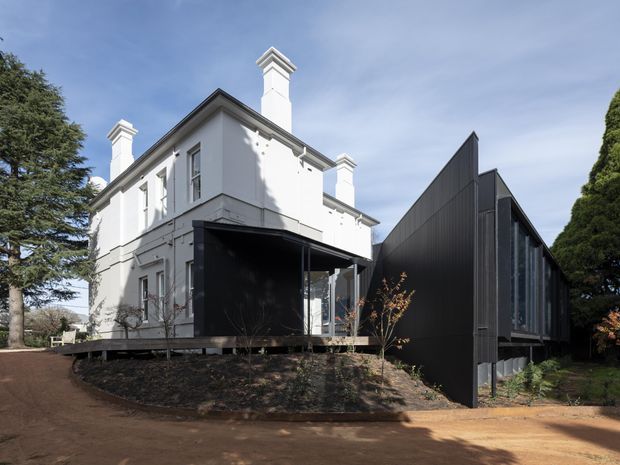 Exterior view of a renovated or extended residence combining a white two-storey heritage-style building with ornate chimney caps and traditional window proportions, alongside a sharply angled dark metal-clad contemporary extension. The buildings sit on a landscaped, slightly sloping site with mature trees and a broad earthen driveway.