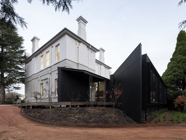 Exterior view of a traditional white rendered multi-storey house with ornate chimney caps, connected to a sharply contrasting contemporary black-clad extension with a steep angular roofline and extensive dark glazing. The building sits within a landscaped garden setting with mature trees and a curved dirt driveway or path.