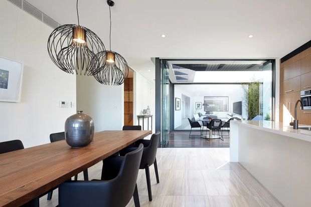 Bright contemporary open-plan dining and kitchen interior with a long dark timber dining table, black upholstered dining chairs, sculptural black wire pendant lights, white walls, light timber flooring, and streamlined white-and-timber kitchen cabinetry. Large open glazing and a visually connected living area create an airy, indoor-outdoor feel.