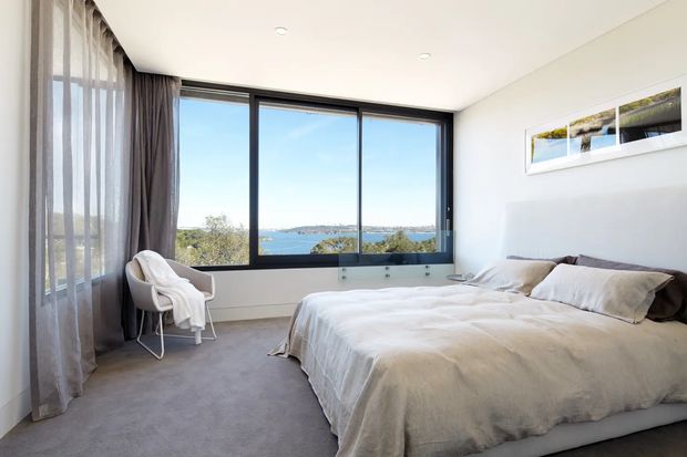 Bright, serene contemporary bedroom with a large floor-to-ceiling corner window overlooking a coastal bay. The room features a low-profile upholstered bed with white bedding, a white sculptural lounge chair, sheer and dark grey curtains, light walls, recessed ceiling lights, and grey carpet. A narrow framed landscape artwork hangs above the bed.