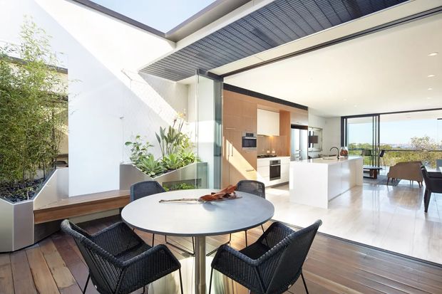 Bright contemporary open-plan kitchen and dining area opening onto a planted outdoor terrace. The room features a white kitchen island and cabinetry, warm timber joinery, large glazed openings, pale polished flooring, and a round dining table with dark woven chairs. A slatted overhead exterior screen and built-in planter add architectural interest, with distant views visible through the glazing.