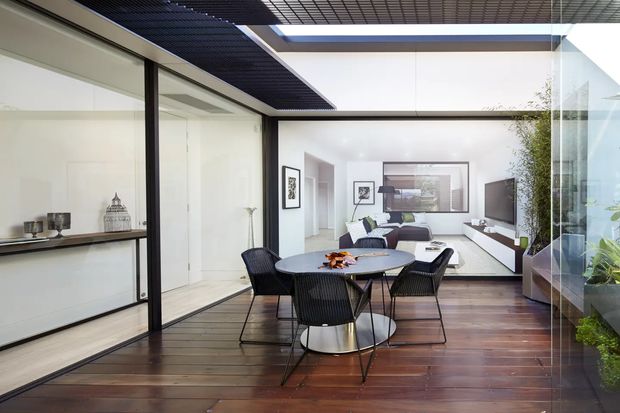 Bright contemporary open-plan interior centred on a dining area, with a dark timber floor, round dining table and black woven chairs. Full-height black-framed glazing and glass partitions surround the space, opening visually to a minimalist living room with a wall-mounted television, low joinery, artwork and pale furnishings. A gridded dark ceiling and indoor planting add architectural and biophilic detail.