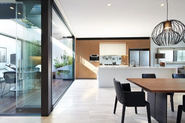 Bright open-plan kitchen and dining area with a contemporary minimalist fit-out. The room features pale timber flooring, extensive black-framed floor-to-ceiling glazing, a timber veneer kitchen wall with integrated appliances, a white island bench, and a timber dining table with dark upholstered chairs. A planted glazed courtyard or sunroom is visible beyond the glazing, and a sculptural black pendant light hangs over the dining area.