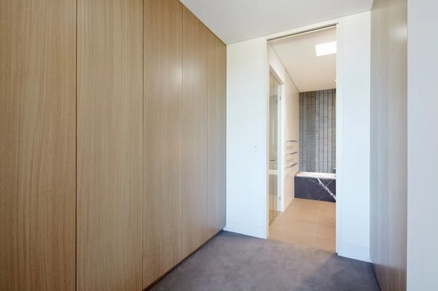 A bright hallway or landing with floor-to-ceiling light timber built-in wardrobe cabinetry on the left and grey carpet underfoot. An open doorway leads directly into a modern bathroom with beige floor tiles, a freestanding or built-in bath, grey tiled walls, and white cabinetry. The space has clean, contemporary detailing and a neutral palette.