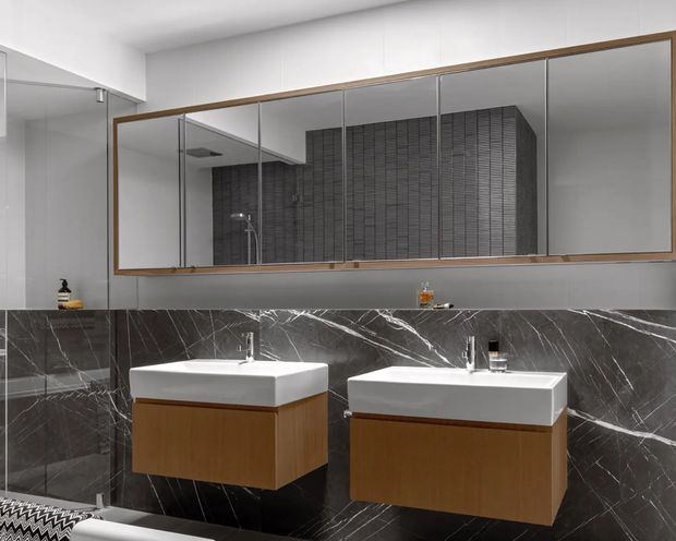 A contemporary double-vanity bathroom with two wall-mounted timber-front basins, white rectangular countertop basins, and a large horizontal mirrored cabinet framed in timber. Dark grey marble-look wall tiling with prominent white veining wraps the vanity wall and shower area. A frameless glass shower enclosure is visible at left, with a dark textured feature tile reflected in the mirror. The room has a clean, refined, high-contrast palette of white, dark grey, and warm wood.