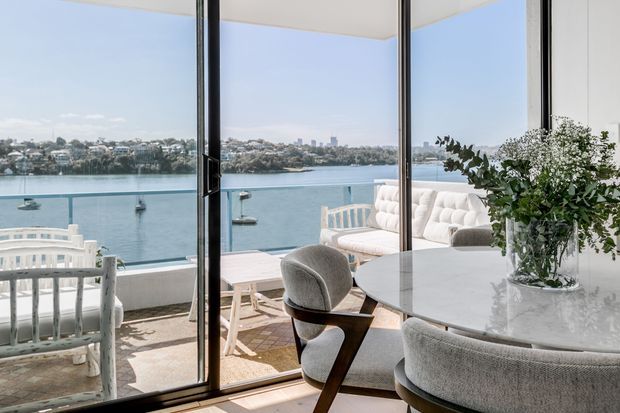 Bright, sophisticated dining and living area with floor-to-ceiling glazing overlooking a calm waterfront. The room features a round marble dining table, upholstered curved dining chairs, a white tufted sofa, pale timber and woven furnishings, and a balcony with additional seating. The interior has a refined contemporary coastal feel with abundant natural light and expansive harbour views.