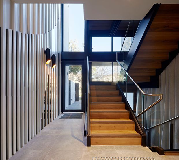 Contemporary residential entry hall with a timber staircase rising to the right, glass balustrades, dark-framed glazing, and a vertical timber slat feature wall. Warm timber stair treads contrast with pale large-format floor tiles and dark metal framing. Abundant natural light enters through tall windows and clerestory glazing, creating a refined, airy circulation space.