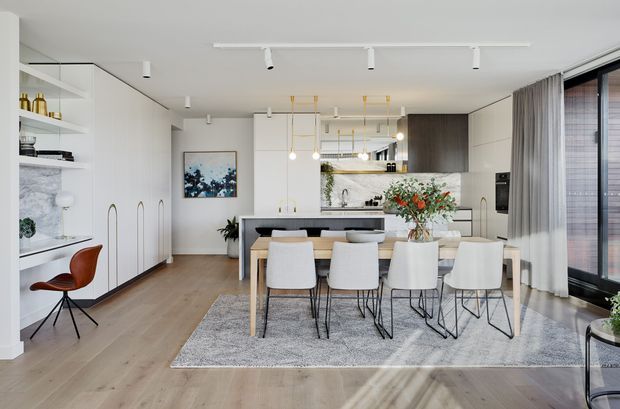 Bright open-plan kitchen and dining area with a large island, white dining table and chairs, light timber flooring, white built-in cabinetry, marble-look shelving, pendant lights, and full-height glazed doors with curtains. The space has a clean, refined contemporary fit-out with warm wood tones and abundant natural light.
