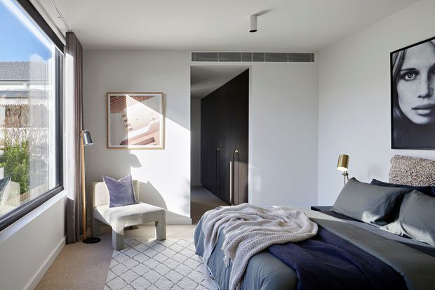 A bright, refined contemporary bedroom with a large floor-to-ceiling window, upholstered armchair, layered bedding, bedside table and wall-mounted artwork. The room features a dark built-in wardrobe or doorway, neutral white walls, soft grey-blue textiles, warm timber-toned accents and a patterned rug. Strong daylight creates an airy, calm atmosphere.
