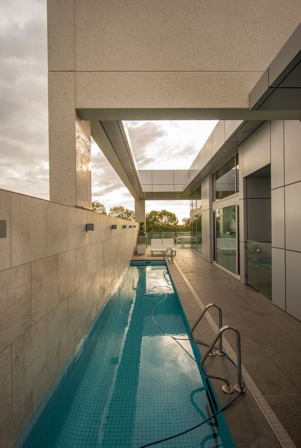 A narrow tiled swimming pool runs alongside a modern building in a partially enclosed outdoor courtyard or terrace. The space features large-format stone or concrete wall and ceiling panels, sleek dark-framed glazing, pale metal facade panels, architectural overhangs, pool ladders, and a linear drainage channel. Trees and an overcast sky are visible beyond the open end.