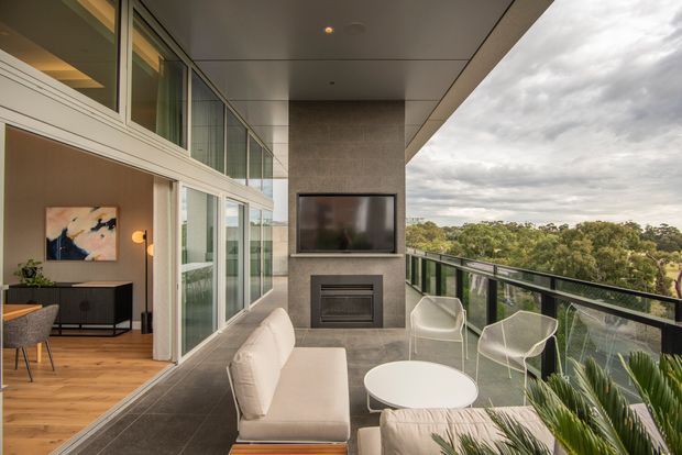 A contemporary covered balcony or terrace overlooking a leafy landscape, furnished as an outdoor lounge with a white sofa, round coffee table and wire-frame lounge chairs. A built-in fireplace and wall-mounted television form a central feature wall, while full-height glazed sliding doors open to an interior dining or living area with timber flooring and a dark console. The space has glass balustrades, warm neutral finishes and an expansive covered ceiling.