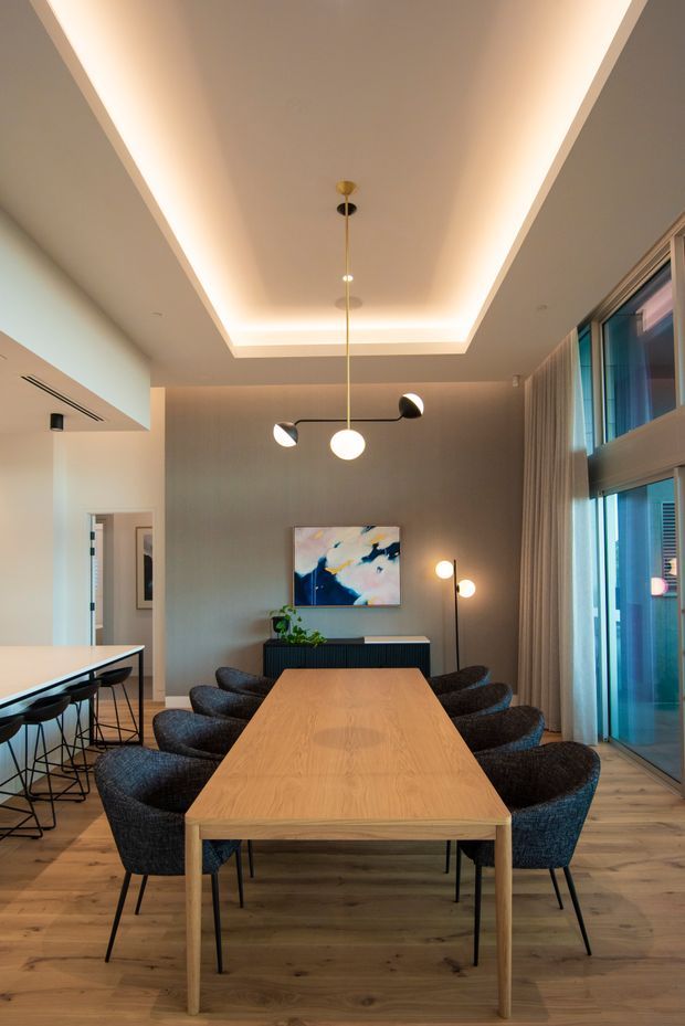 A contemporary dining room centred on a long light-oak dining table surrounded by dark upholstered chairs. The room features timber flooring, a textured grey feature wall, a linear black sideboard, abstract artwork, floor-to-ceiling glazed doors with curtains, and sculptural pendant and floor lighting. A recessed tray ceiling with warm concealed LED lighting creates a refined, inviting atmosphere. A kitchen or breakfast counter is partially visible through the open area on the left.