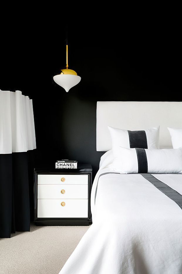 A refined bedroom interior with a bold black accent wall, crisp white upholstered bed and bedding, and black-and-white striped detailing. A black bedside table with white drawers and brass pulls sits beside the bed, complemented by a sculptural pendant light with a gold finish and white shade. White curtains with black drapery and light neutral carpeting complete the high-contrast scheme.