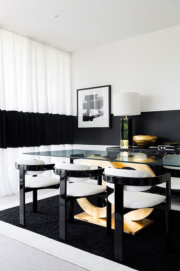 A bright, high-contrast dining room with a large rectangular glass dining table surrounded by black-framed chairs with white upholstered seats. The room features white walls and sheer white curtains contrasted by a broad black wall panel, a sculptural gold-toned table base, black textured rug, framed monochrome artwork, and a table lamp with a reflective gold-and-black base. The styling is polished and dramatic with strong geometric forms.
