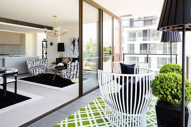 Bright contemporary apartment living area opening onto a balcony through full-height glazed sliding doors. The interior features a compact kitchen, black-and-white patterned lounge chairs, a black rug, sculptural chandelier, and modern artwork. The balcony has a striking white sculptural chair, green-and-white patterned rug, potted topiary, and views toward neighbouring apartment buildings.