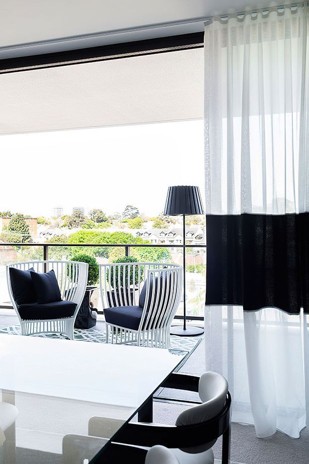 Bright contemporary living and dining area with floor-to-ceiling glazing opening onto a balcony. The room features black-and-white furnishings, a white dining table, sculptural black dining chairs, two white-framed lounge chairs with dark cushions, sheer white curtains with a black band, and a slim black floor lamp. A leafy suburban outlook is visible beyond the balcony.
