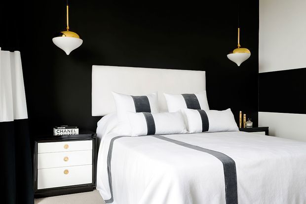 A high-contrast bedroom featuring a white upholstered bed with crisp white bedding accented by dark charcoal-grey stripes, set against a predominantly black wall. Matching black-and-white bedside furniture and pendant lights reinforce the monochromatic scheme, with a small Chanel decorative item on the bedside table.
