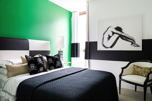 A bright, contemporary bedroom featuring a large vivid green feature wall, a black-and-white upholstered bedhead, layered white bedding with a dark textured bedspread, and skull-patterned accent cushions. Black-and-white window curtains, monochrome artwork, and a dark-framed upholstered armchair complete the bold graphic scheme.