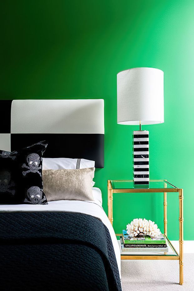A close-up of a boldly styled bedroom corner with a vivid emerald green wall, a partially visible bed with a black upholstered headboard, white bedding, black textured coverlet, and decorative black-and-white pillows. A glass-topped brass side table holds a striped black-and-white table lamp, books, and a small white floral arrangement. The room has strong graphic contrast and a polished, dramatic look.