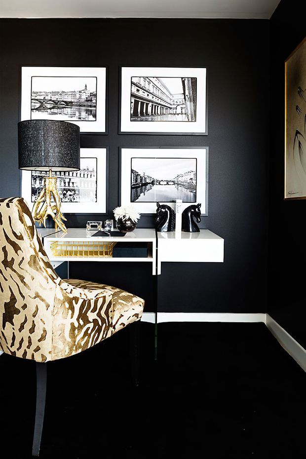 A dramatically styled interior corner with black walls and glossy black flooring, featuring a white floating console desk, black-and-white framed architectural prints, a sculptural gold-based table lamp, decorative chess pieces, and a patterned upholstered armchair. The space has a high-contrast, refined, art-focused atmosphere.