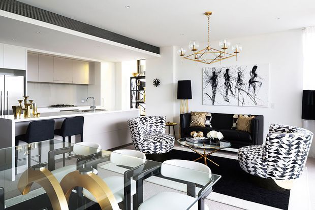 Bright open-plan contemporary living and dining area with a white kitchen, island bench and bar seating, a glass dining table with white chairs, and a black sofa arranged around a glass coffee table. Geometric black-and-white patterned armchairs, monochrome artwork, brass geometric pendant lighting, and black accents create a refined graphic interior.