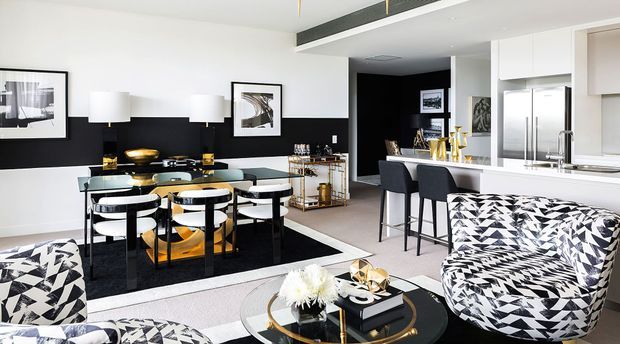Open-plan contemporary living, dining and kitchen area with a bold black-and-white palette. The space features a glass dining table with upholstered chairs, patterned lounge seating, a black feature wall, white cabinetry, stone-look benchtops, bar seating and metallic accents.