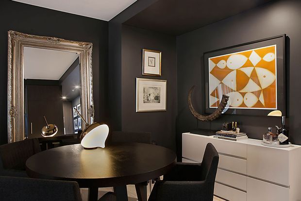A moody, refined dining room with charcoal walls and ceiling, a round dark dining table, upholstered dining chairs, and a white contemporary sideboard. A large ornate wall mirror, framed artwork, sculptural wall decor, books, and decorative objects add visual interest. The room appears compact with standard-height ceilings and a strong black, white, and warm ochre palette.
