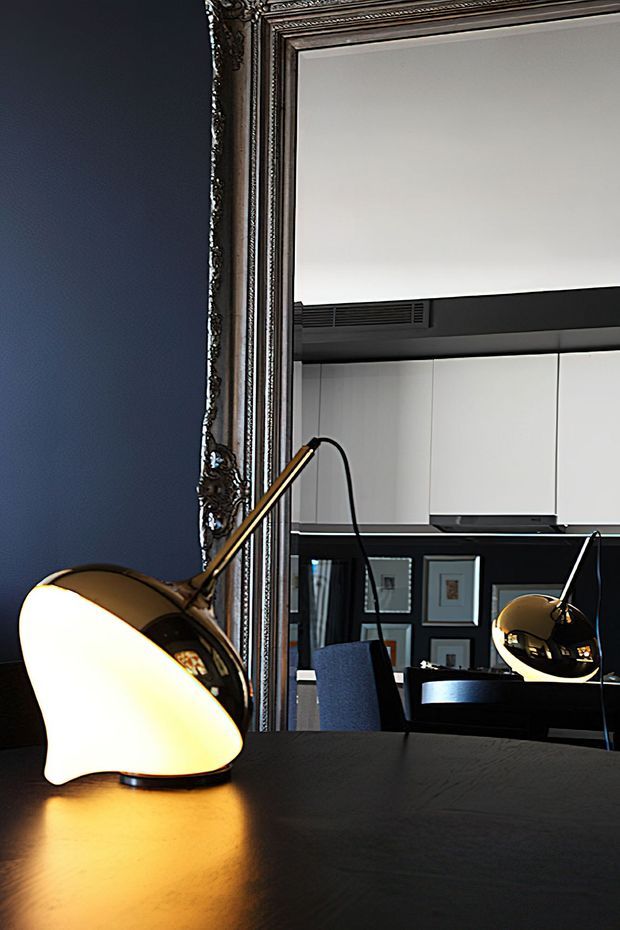 A moody interior detail featuring a dark blue wall, an ornate antique-style framed mirror, and a glowing brass desk lamp on a dark tabletop. The mirror reflects a sleek contemporary kitchen or workspace with white cabinetry, black accents, framed artwork, and a second lamp, creating a striking blend of classic decorative detailing and modern furnishings.