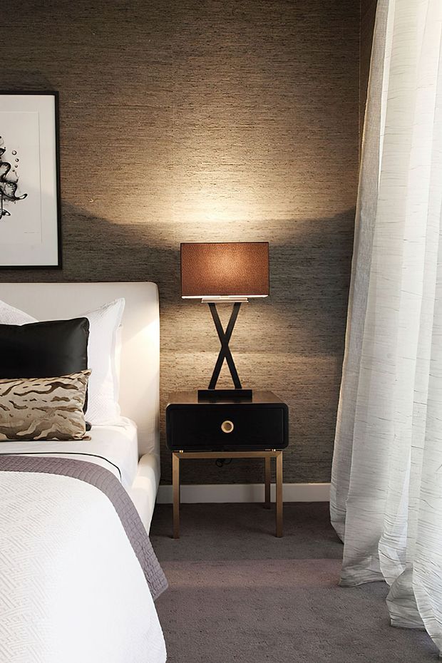 A close-up view of a refined bedroom corner featuring a partially visible upholstered bed with crisp white bedding, layered neutral and patterned pillows, a black bedside table with slender metallic legs, and a sculptural black table lamp with a warm brown shade. The dark textured feature wall contrasts with sheer white curtains and soft grey carpet, creating a warm, sophisticated atmosphere.